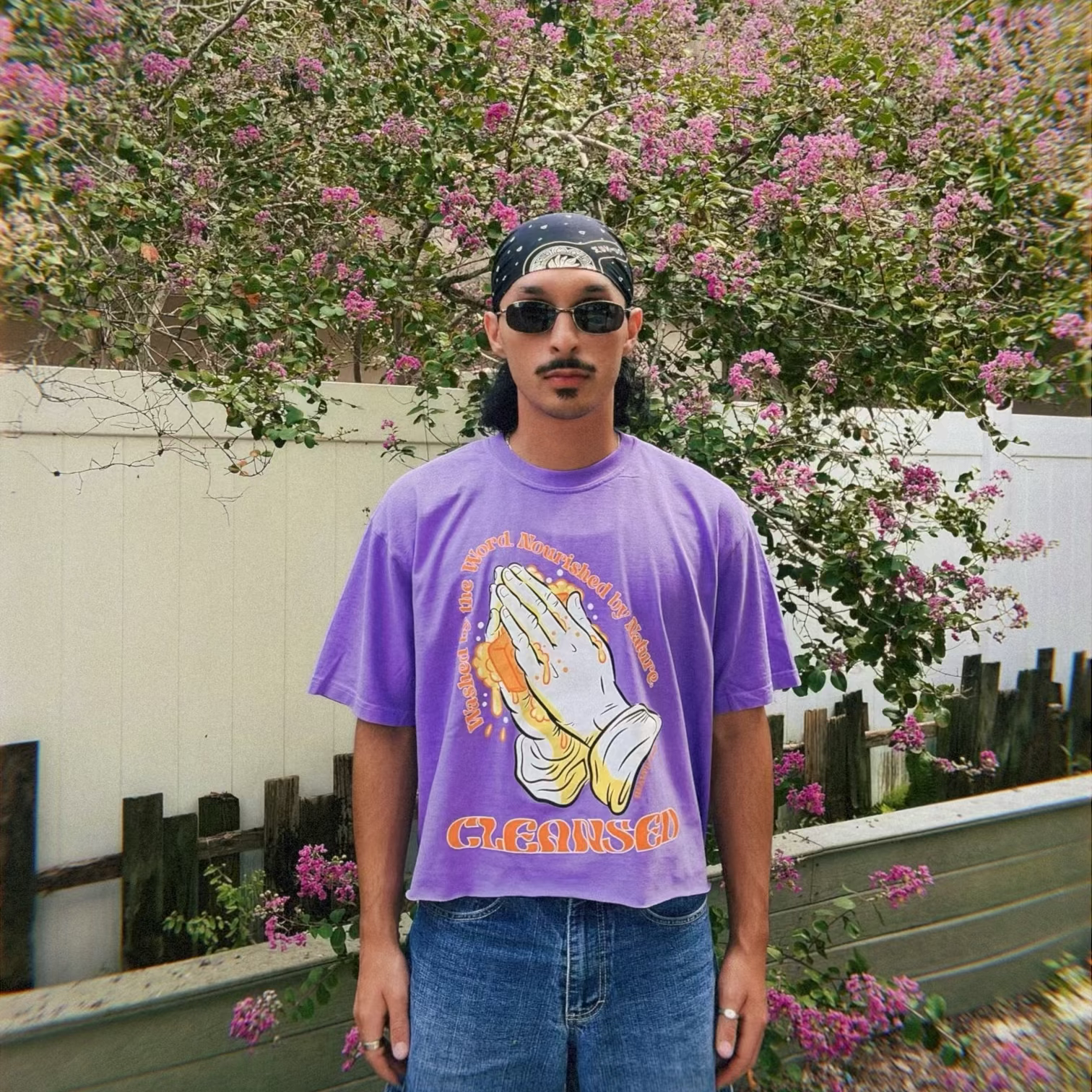 Person wearing a purple t-shirt with a graphic design, standing in front of a flowering tree.